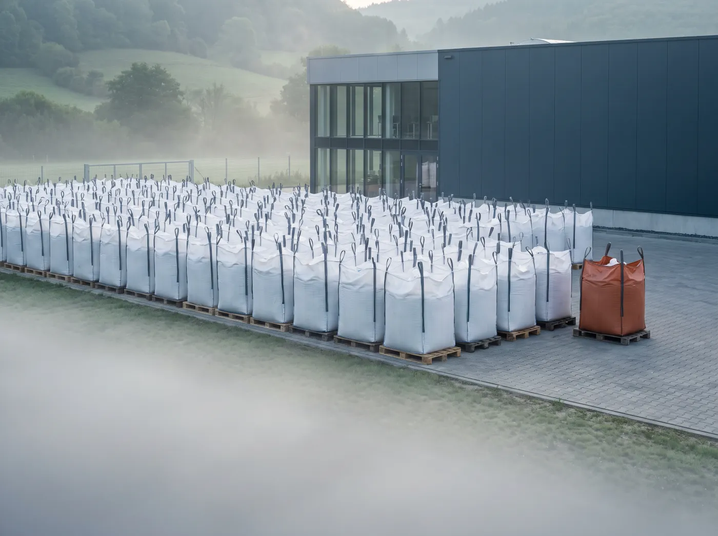White bulk bags in rows outside a factory, one orange bag held apart from the rest